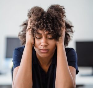Woman in office setting expresses stress, seated at desk with hands in hair. Perfect for workplace stress concept.